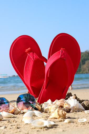 Stock photo showing close-up view of tinted, mirrored sunglasses besides a pile of seashells in front of a pair of red flip flops standing up, forming a heart shape, in the sand on a sunny, golden beach with sea at low tide in the background. Romantic holiday and honeymoon concept.