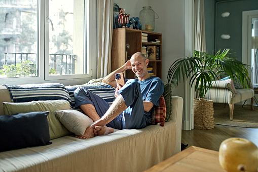 Full length view of casually dressed man in mid 40s reclining in living room, holding smart phone, and laughing while communicating with friend.