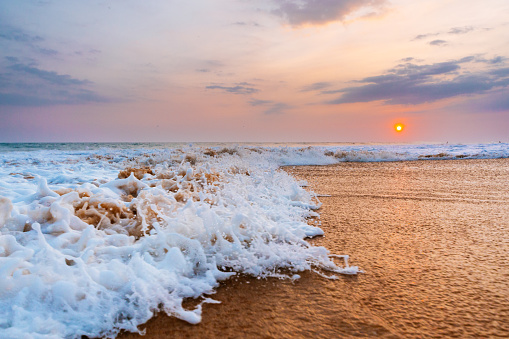 Sunset illuminates the sky with warm hues as ocean waves, captured mid-crash, froth onto a sandy beach. The close-up perspective highlights the dynamic movement of the water. The waves are white and frothy, and the sand on the beach appears wet and reflects some light from the sunset. The sun is visible near the horizon over the calm sea beyond the waves.