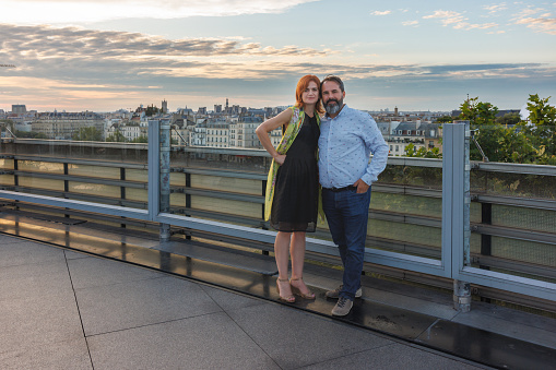 Middle aged couple standing embraced by the fence on top of building in the city and smiling at camera, staycation in Europe, Paris, France
