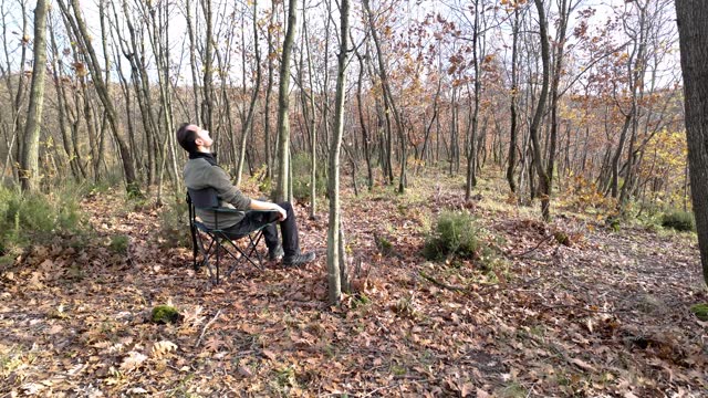 Male camper sets a camping chair and sits on it for relaxing in a forest filled with autumn leaves.