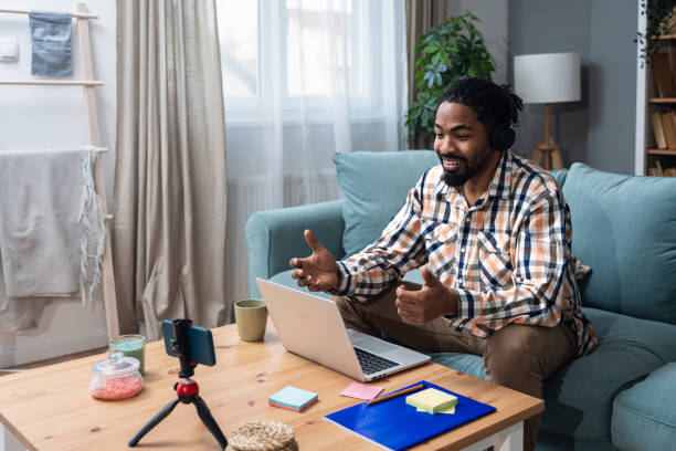 istock Young African American man with headset having web call using laptop computer and smartphone sitting on sofa talking at webcam in home office Video conference online business product strategy meeting 1952109868