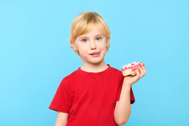 ブロンドの髪とそばかすのあるかわいい男の子が、舌を突き出して、艶をかけられたドーナツを楽しんでいます。子供と甘いジャンクフードのコンセプト。色とりどりのドーナツを持った男� - preschooler caucasian one person part of ストックフォトと画像