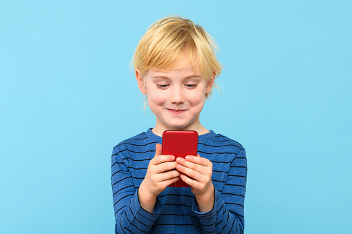 Cute child looking at his smartphone smiling. Boy playing video games on his phone, isolated over pastel blue background. Children and technology.