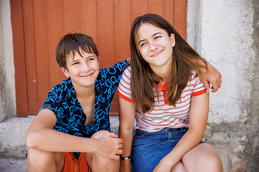 Cute teenage brother and sister sitting on stairs in front of an old house in Croatian insular town, embracing and smiling happily
