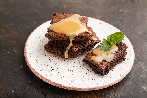 Chocolate brownie with caramel sauce with a cup of coffee on black concrete background. side view, close up.