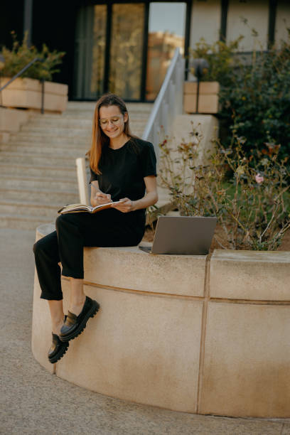 istock Young female student in eyeglasses making notes and browsing laptop outdoors near city building 1952158066