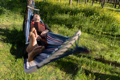 young adult man lying in a hammock held between trees, while sleeping with a book on top