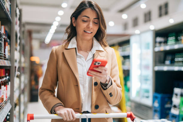 istock Smiling woman pushing trolley in supermarket while checking messages 1952203878