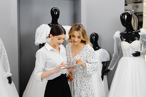 Beautiful bride discussing details with the wedding planner in store. Attractive bridal shop owner women use digital tablet help customer choosing wedding gown at the store.