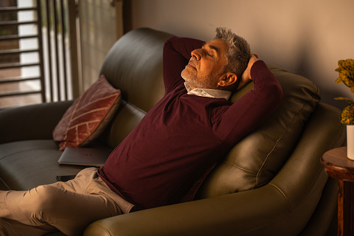 High angle view of senior man with hands behind head and eyes closed relaxing on cozy sofa in living room at home