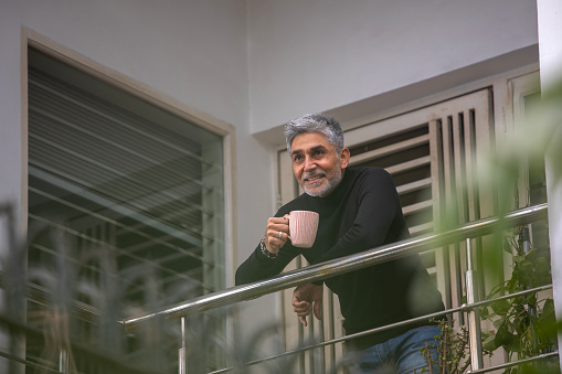 Low angle view of smiling handsome senior man with coffee mug looking away thoughtfully and standing by railing in balcony