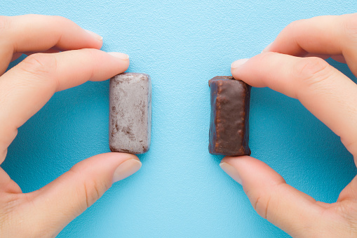 Young adult woman hand fingers holding fresh and old spoiled dark chocolate candies on light blue table background. Pastel color. Compare two pralines. Point of view shot. Top down view.