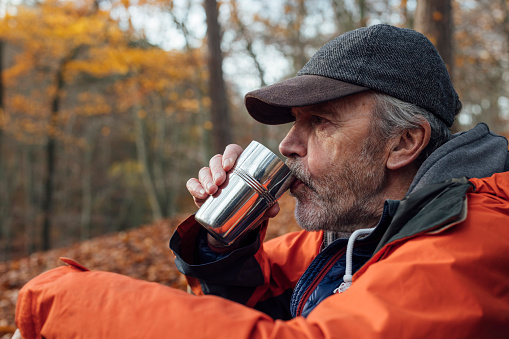 A senior man enjoying a day out in nature at the weekend in a woodland area in Hexham, North East England. He is taking a break from walking and enjoying a hot drink from a thermal cup while sitting on the ground.

Videos available that are similar to this scenario.