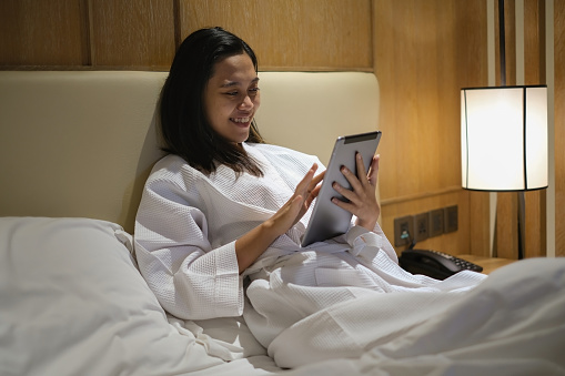 Close-up shot of Indonesian young woman using hotel tablet technology in the room, to access an array of hotel services at the tap of a touchscreen.  Concept : single female traveller, watching a movie, reading a book, surfing the internet, looking at social media / viral video.