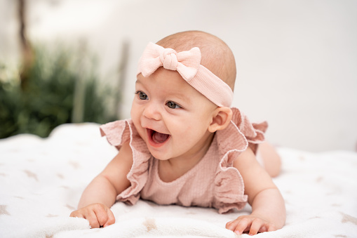Close-up portrait of a little baby laughing with her mouth open dressed in a ruffled shirt and a pink bow on her head lying on a blanket.