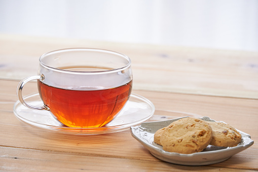 Part view of a cup of golden tea, with a home-baked almond biscuit on the saucer. White porcelain cup and saucer on a rustic wooden table surface with a white teapot in the background. Outdoor shot with natural light.