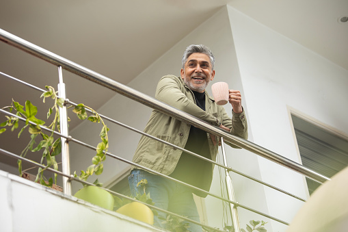 Low angle view of smiling handsome senior man with coffee mug looking away thoughtfully and standing by railing in balcony