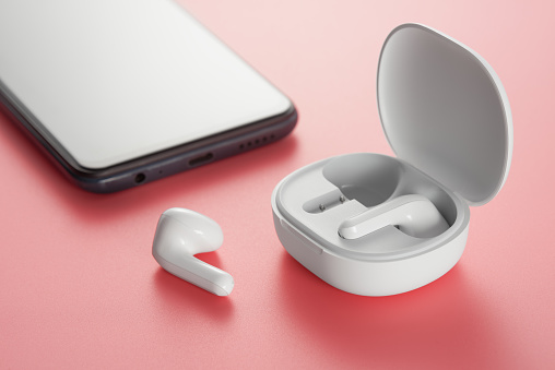 White wireless headphones in a charging case and a smartphone on a pink background.