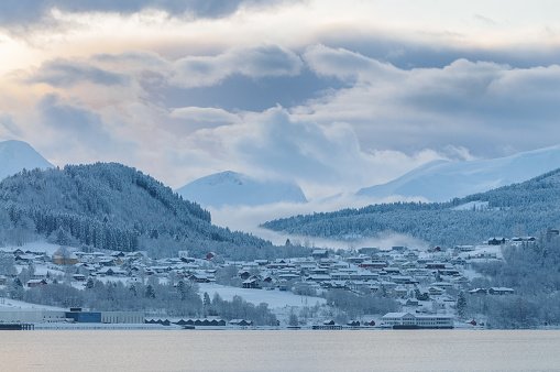 A serene snowy landscape showcases a quiet village beside the sea, enveloped by gentle mist and surrounded by forested mountains at dawn.