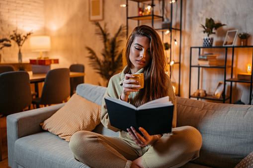 Beautiful young woman reading a book and drinking coffee at home at night.