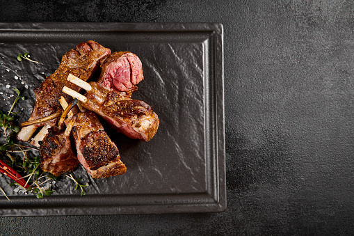 Juicy grilled lamb rack with a crusty sear, accented by fresh green herbs and a fiery red chili on a dark slate plate.