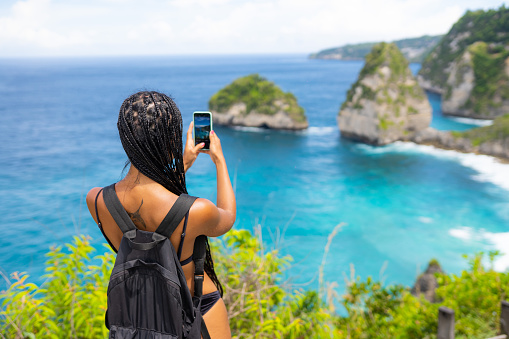Brunette woman taking pictures at Kelingking beach. Blues and green sand and the beautiful mountain shaped on the sea.Travel destinations vacations people sharing social media concept nature scenics