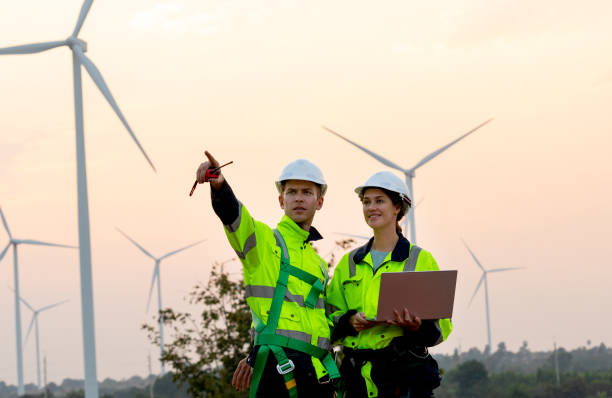 istock Technician workers use laptop to work and stay with her co-worker point to left side in evening with windmill or wind turbine on the background. 1952442128