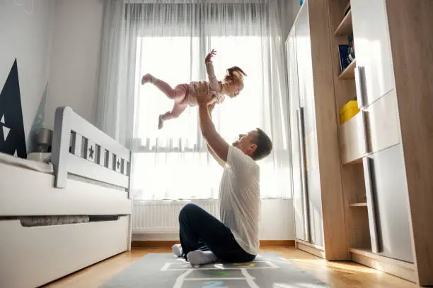 A fun dad is playing with his little girl while tosing her in the air in nursery room.
