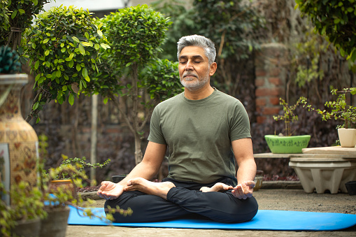 Full length of senior man meditating in lotus position while sitting on exercise mat surrounded with plants in park