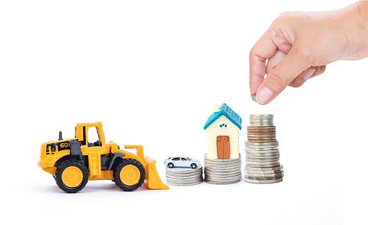 Miniature house and car model on coin stack with hand holding coin with front loader truck on white texture background, property investment, real estate business, buy new house