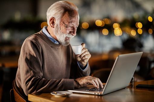 Portrait of a senior casual businessman sitting in cafe, typing on a laptop and smiling at it while drinking coffee.