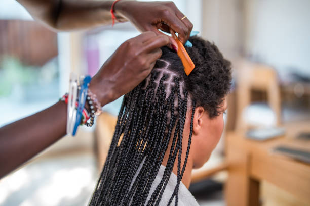 A mid adult Black woman braids the hair of a mixed race female in a domestic setting, showcasing a traditional hairstyle being made.