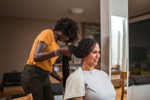 Mid adult Black female hairstylist professionally braiding the hair of a Mixed race client in a cozy home setting.