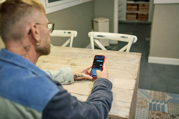 Bearded man sitting at kitchen table and text messaging Over the shoulder view with focus on smart phone as individual in early 50s communicates via smart phone. man looking at phone over shoulder stock pictures, royalty-free photos & images