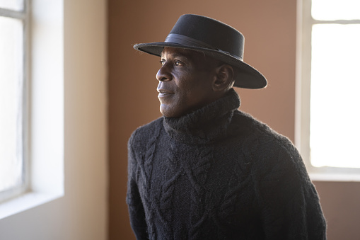 A senior African-American man is standing by a bright sun-lit window, wistfully looking outside, dressed in a jumper and hat ready to go outside