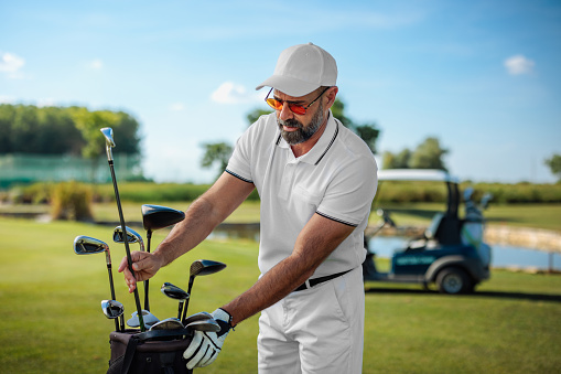 Caucasian mature golfer standing on a golf field, choosing a correct golf club from his golf bag