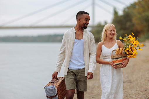 An multiethnic happy couple walking alongside the river, carrying picnic supplies and smiling