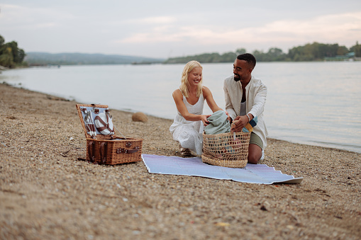 Couple is kneeling on a blanket, smiling before preparing for a picnic by the river during a hot summer day