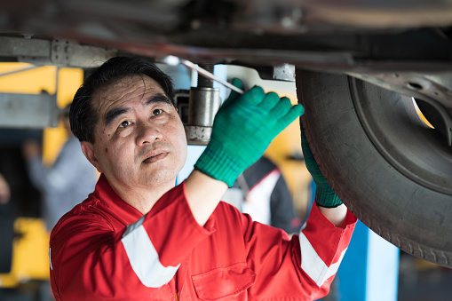 Male car mechanic worker working underneath lifted car. Asian male mechanic vehicle service maintenance checking under car condition in garage. Auto car repair service and maintenance concept