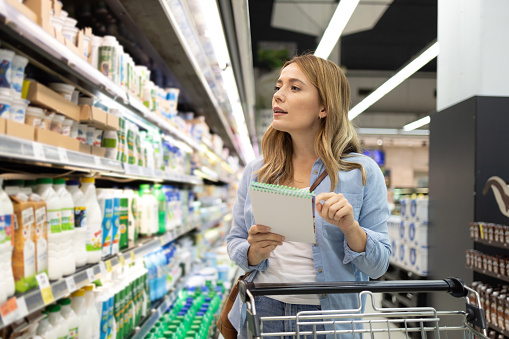 A young housewife holding her shopping list in her hands is looking at the diary products in a supermarket