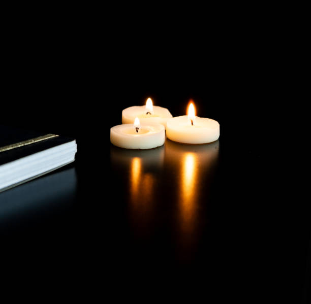 istock Closed notebook on a black table with 3 small lit tea light candles and their reflection - black background and empty space 1952656536