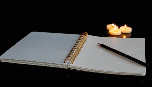 Open spiral notebook on a white page with a pencil on a black table with three small lit candles