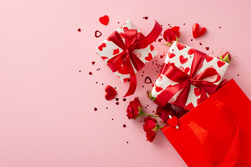 Celebration surprise: paper bag with gift boxes, adorned in themed paper, red roses, and heart-shaped confetti. Top view on pastel pink backdrop, leaving space for a message or promotion