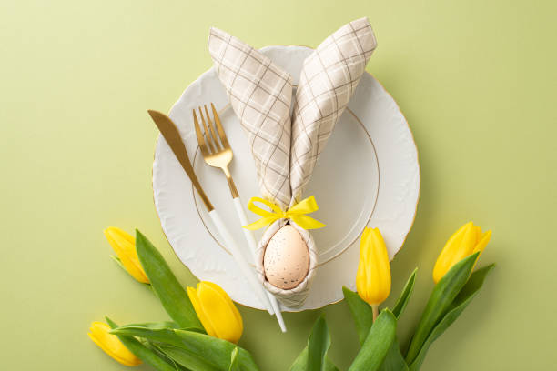 istock Eggstra Special Table: Top-down view of an enchanting Easter arrangement featuring a plate, bunny ears napkin, cutlery, bouquet of fresh yellow tulips. Pastel green background with space for text 1952672321