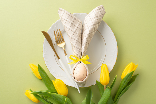 Eggstra Special Table: Top-down view of an enchanting Easter arrangement featuring a plate, bunny ears napkin, cutlery, bouquet of fresh yellow tulips. Pastel green background with space for text