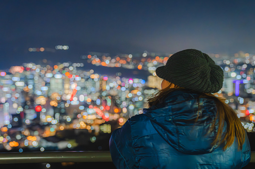 The image of a young woman standing and admiring the night view of Seoul at an observation deck on Namsan Mountain.