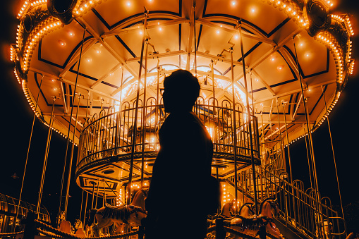 The silhouette of a young man who is standing vacantly Under the lights of the carousel at night.