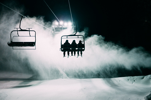 Family skiing at night sit on ski lift while snow cannons are in operation