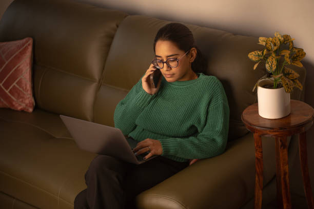 woman with mobile phone and laptop working at home - multi tasking asian and indian ethnicities asian ethnicity lifestyles imagens e fotografias de stock